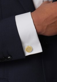 Gold-tone cufflink with a textured, circular surface, attached to a white shirt cuff, contrasted against a dark blue suit jacket.