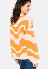Oranje en crèmekleurige oversized trui met golvend patroon, gebreide textuur en geribde manchetten. Achteraan is de ontspannen pasvorm zichtbaar.
