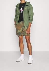 Zelená lehká bunda s kapucí, černé tričko s bílou grafikou, maskáčové šortky a bílé tenisky. Ležérní letní outfit.