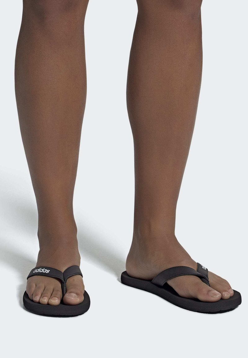 Chanclas negras con una plantilla texturizada; una correa estrecha presenta el logo de adidas. La suela es plana y flexible, diseñada para la comodidad.