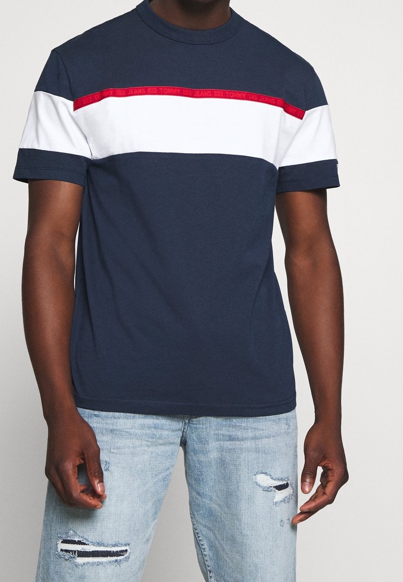 Muž ve navy modrém a bílém tričku s barevným blokem, s červeným pruhem Tommy Jeans, zkombinovaným se světle modrými roztrhanými džíny.