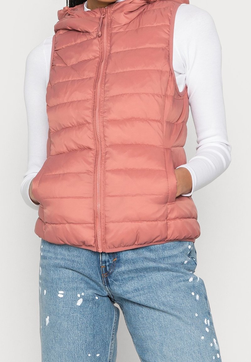 Femme portant un gilet sans manches matelassé rose poudré sur une chemise blanche à manches longues et un jean bleu éclaboussé de peinture, les mains dans les poches du gilet.