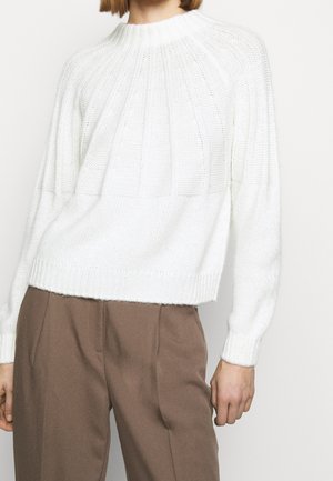 Femme portant un pull en maille côtelée blanche associé à un pantalon marron taille haute, montrée du cou jusqu'aux hanches.