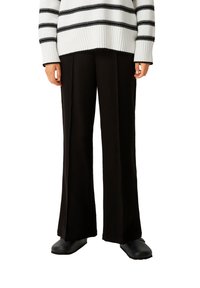 Schwarze weit geschnittene Hose, kombiniert mit einem oversized Sweatshirt in Weiß und Schwarz gestreift, das gerippte Bündchen und eine lockere Passform aufweist. Schlichte schwarze Schuhe.