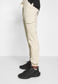 Pantalons cargo beige avec de multiples poches, dotés d'une taille élastique avec cordon de serrage et de bas de pantalon fuselés. Portés avec des chaussures de sport noires.