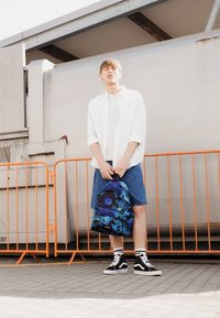 Niebieski plecak w kwiatowy wzór, biała koszula z guzikami, jasny t-shirt, dżinsowe szorty i czarne wysokie trampki. Tło z betonem.