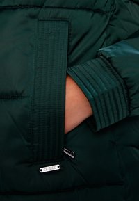 Giacca imbottita verde scuro con polsini a coste, tasca esterna e placca del logo Guess argento vicino all'orlo. Tessuto liscio e isolato.