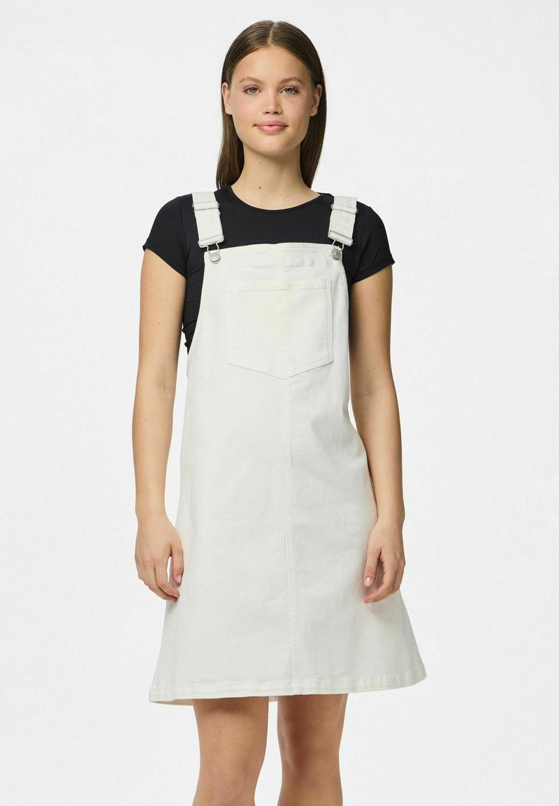 Bela denim igralna obleka z nastavljivimi naramnicami, sprednjim žepom in širokim krojem. V kombinaciji z črno majico s kratkimi rokavi spodaj.