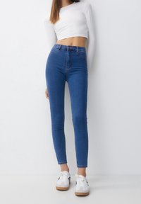 Mujer vestida con jeans ajustados de tiro alto azules, una blusa blanca de manga larga recortada y zapatillas blancas, apoyada contra un fondo blanco liso.