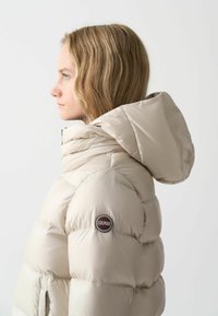 Beige puffjacka med huva, med en texturerad, vadderad design och ett framträdande Colmar-logotypmärke på ärmen.