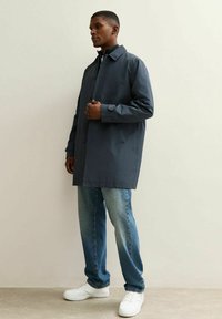 Hombre de pie vistiendo un abrigo abotonado gris oscuro, jeans azules y zapatillas blancas, contra un fondo claro y liso.