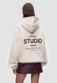 Felpa color crema con un grande cappuccio e polsini a costine, che presenta un testo di un marrone scuro "WRSTBHVR STUDIO GARMENTS" e un piccolo dettaglio del logo.