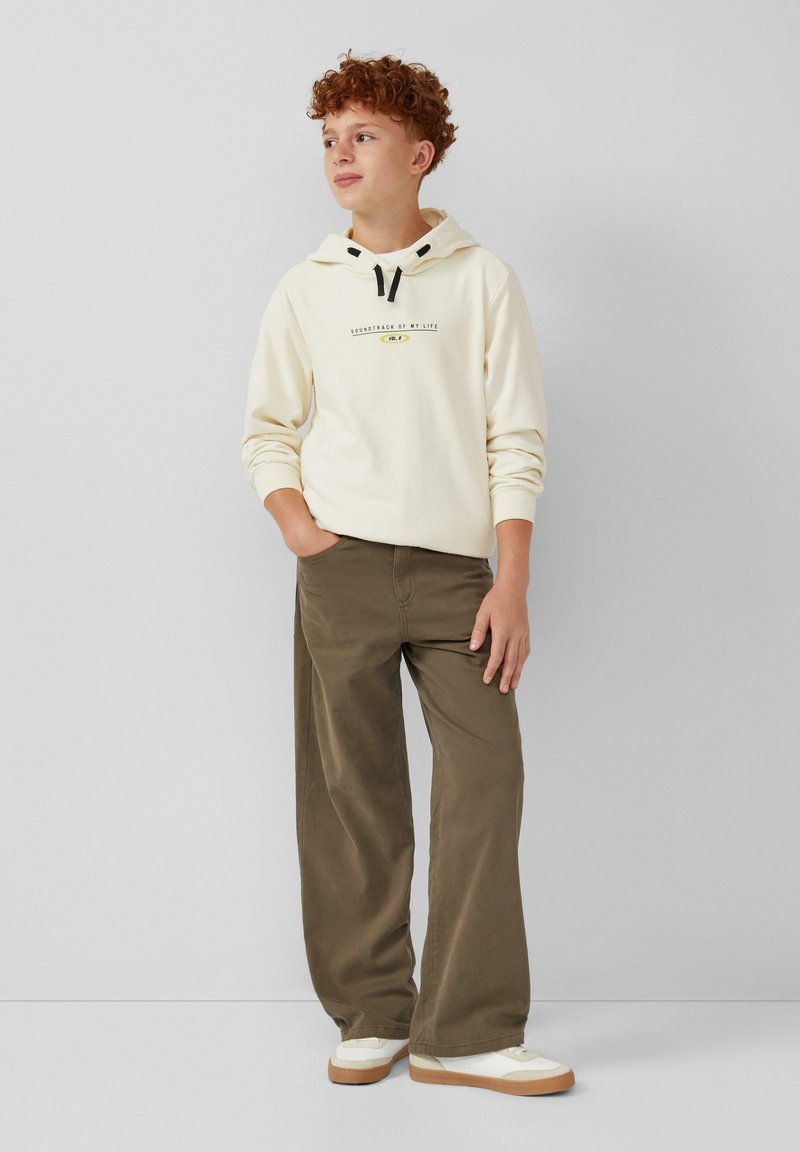 Crèmewitte hoodie met zwarte trekkoord, tekstontwerp; olijfgroene wijde broek; witte schoenen met enkellaarsjes met een tan zool. Eenvoudige, casual outfit.