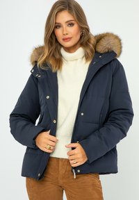 Granatowa kurtka puchowa z kapturem wykończonym futrem, z suwakiem z przodu, zatrzaskami oraz kieszeniami po bokach. Miękka, pikowana tekstura z złotymi akcentami.