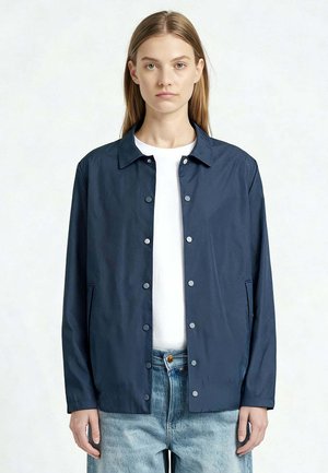 Jong persoon met lang haar draagt een marineblauwe jas met knopen over een wit shirt en lichtblauwe spijkerbroek tegen een effen achtergrond.