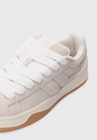 Suede sneakers in light beige with white flat laces. Features a padded collar and cream rubber sole with subtle branding on the side.
