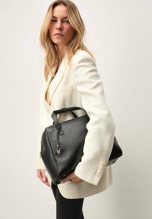 Femme blonde en blazer blanc tenant un grand sac à main en cuir noir avec une fermeture éclair et une poignée dorées, sur un fond uni.