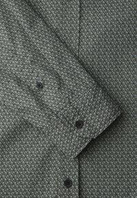 Chemise grise avec motif géométrique de cubes noirs, boutons noirs sur le poignet et la patte de boutonnage avant, manche repliée sur le devant de la chemise.
