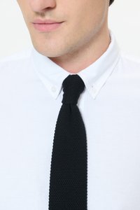 Black knitted tie with a textured finish, worn over a white collared shirt. The tie is slim and features a simple, classic knot.