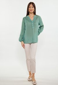 Blusa turchese con maniche lunghe a sbuffo, patta frontale con bottoni e vestibilità rilassata; abbinata a pantaloni slim beige chiaro e ballerine color crema.