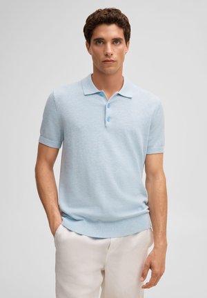 Giovane uomo con capelli castani ricci che indossa una polo azzurro chiaro a maniche corte e pantaloni bianchi, in piedi con una mano in tasca.