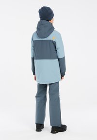 Veste étanche bleu et gris foncé avec capuche, poignets élastiques et détail du logo. Associée à un pantalon assorti et des bottes noires.