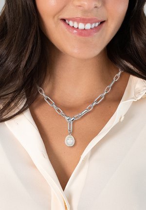 Collana in argento con pendente a forma di perla ovale texturizzata, indossata sopra una blusa di colore chiaro con una lucentezza sottile.