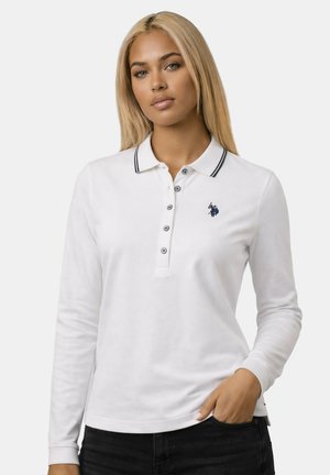 Woman with long blonde hair wearing a white long-sleeve polo shirt with black trim and embroidered logo, hand in pocket.