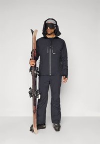 Skiklær i svart, bestående av en vanntett jakke og bukser. Tilbehør inkluderer solbriller, en lue og skistaver med bindinger.