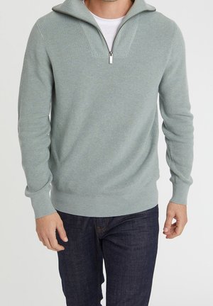 Mann trägt hellgrauen, gerippten Strickpullover mit halbem Reißverschluss am Kragen und dunkelblaue Jeans, stehend vor weißem Hintergrund.