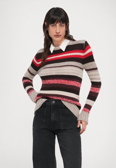 Persona valkā kreklu ar apkakli zem sarkani, melni, bēšā un pelēkā toņos svītrainas adītas džempera, kas kombinēts ar melniem, augsta vidukļa džinsiem.