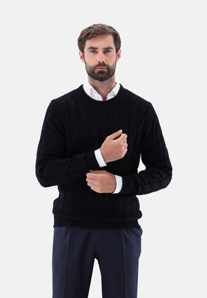 Maglione nero a trecce con scollatura rotonda e polsini a costine, indossato sopra una camicia bianca con colletto, che presenta un aspetto di tessuto strutturato.
