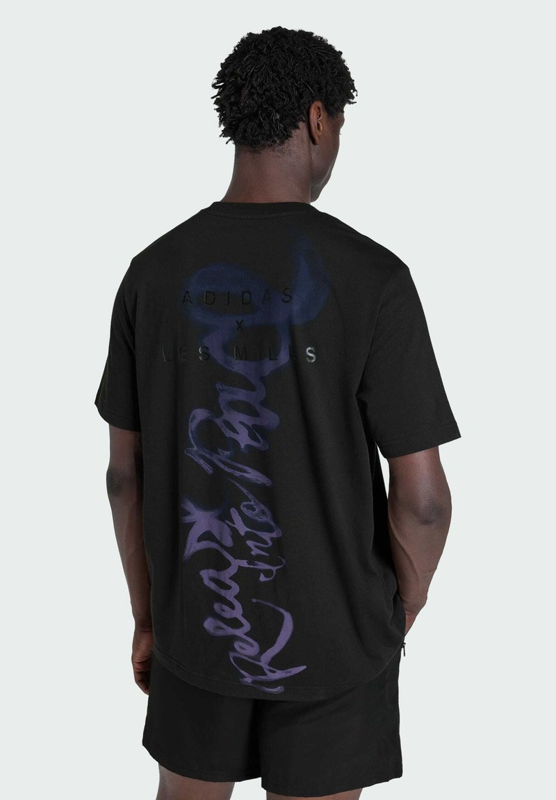 Hombre con cabello corto y rizado que lleva una camiseta negra Adidas x Les Mills con un gran texto vertical en púrpura en la espalda, combinada con pantalones cortos negros.