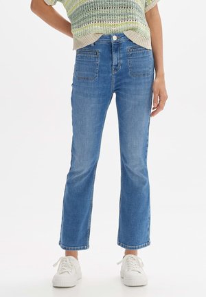 Vrouw draagt een blauwe jeans met hoge taille en rechte pijpen, voorzien van voorzakken, een licht gebreide top en witte platformsneakers.