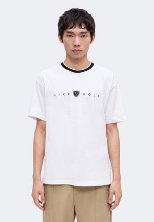Jonge man draagt witte Nike Golf t-shirt met zwarte kraag en beige broek, staand tegen een effen lichte achtergrond.