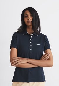 Μπλουζάκι πόλος σε ναυτικό μπλε, με κοντά μανίκια, γιακά και λευκές λεπτομέρειες κουμπιών. Διαθέτει ένα διακριτικό λογότυπο στο στήθος και υφασμάτινη υφή.