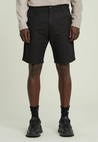 Pantaloni corti neri in cotone con vestibilità rilassata, dotati di chiusura con bottone, tasche frontali e orli rifiniti. Indossati con sneakers nere e calze.