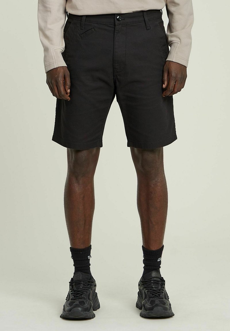 Pantaloni corti neri in cotone con vestibilità rilassata, dotati di chiusura con bottone, tasche frontali e orli rifiniti. Indossati con sneakers nere e calze.