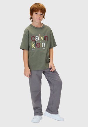Maglietta verde oliva con scritta "calvin klein" multicolore, abbinata a pantaloni della tuta grigi e scarpe bianche. Stile casual, vestibilità comoda.