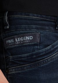 Los jeans de mezclilla azul oscuro presentan una cinturilla texturizada y un bolsillo cosido. Una etiqueta dice "PME LEGEND AMERICAN CLASSIC" en letras blancas.