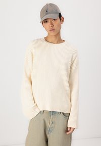 Lys beige ribbestrikket genser med rund hals og vide ermer, kombinert med lyseblå denimjeans og en grå caps.