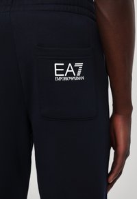 Tengerészkék melegítőnadrág hátsó zsebbel, amelyen a fehér EA7 Emporio Armani logó található. Az anyag lágy és sima tapintású.