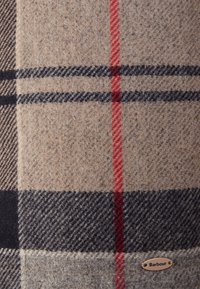 Wełniany koc w kratę w odcieniach beżu, czerni i czerwieni, z teksturowaną powierzchnią oraz metalową metką oznaczoną "Barbour" na krawędzi.