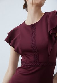 Burgundy klänning med volangärmar, med texturerad spetsdetalj längs framsidan och bälte i midjan, tillverkad av slätt tyg.