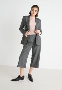 Grijze geblokte blazer over een roze geribbelde top, gecombineerd met brede grijze broek en zwarte enkelboots met een lage hak.