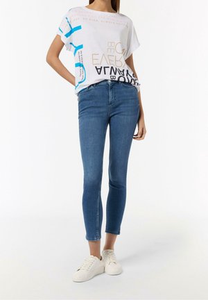 Femme portant un t-shirt blanc graphique, un jean skinny bleu et des baskets blanches, debout avec une main dans la poche sur fond blanc.