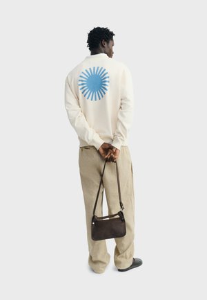 Person standing facing away wearing cream sweatshirt with blue sun graphic, beige pants, holding small dark brown handbag behind their back.