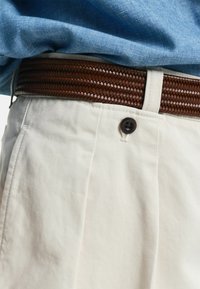 Un cinturón de cuero marrón trenzado contrasta con unos pantalones de trenza beige claros, que presentan un botón marrón y una superficie texturizada para añadir detalles.