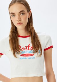 Hvid ribbet crop top med rød kant, der har blå og rød tekst: "Boston Massachusetts new league athletic district." Glat tekstur.