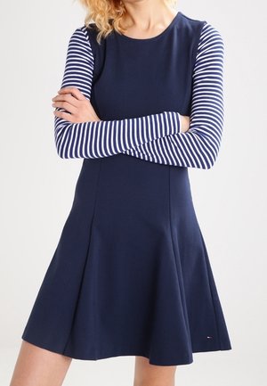 Mujer vistiendo un vestido azul marino con corsé ajustado y falda acampanada, superpuesto sobre una camisa de rayas azul marino y blanca de manga larga, con los brazos cruzados.